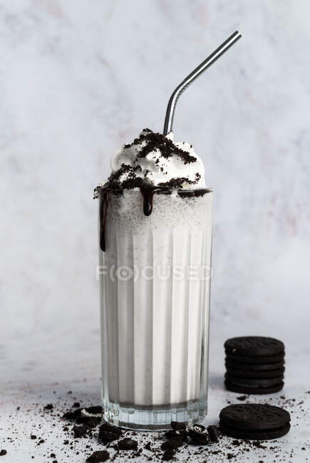 Vaso de batido dulce blanco rematado con crema batida y migas de galletas de chocolate servidas con paja en una mesa desordenada cerca de la pared - foto de stock