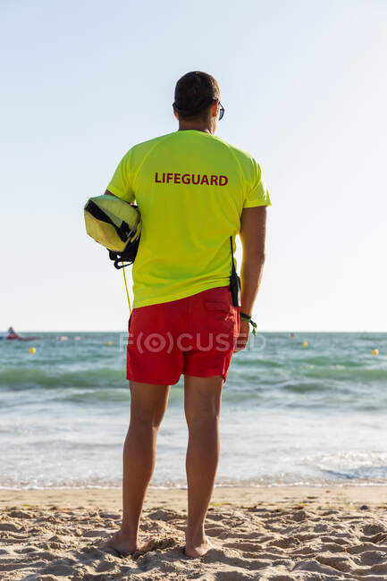 Full body back view of anonymous male lifeguard in shorts and t shirt and maintaining safety on sandy seashore — Stock Photo