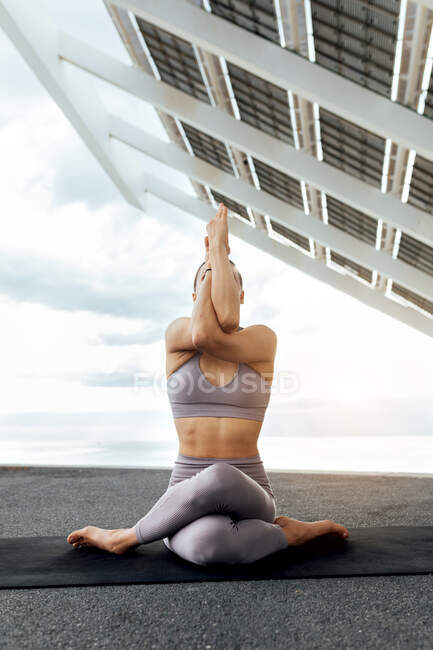 Corpo inteiro de fêmea anônima sem rosto magro em activewear fazendo Gomukhasana com braços de águia enquanto sentado perto do painel solar na rua — Fotografia de Stock