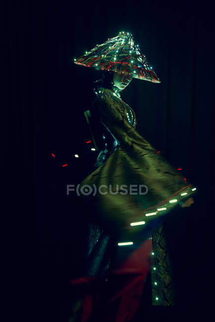 Anonymous female in authentic outfit and Vietnamese hat with glowing illumination standing in dark modern studio on black background during performance — Stock Photo