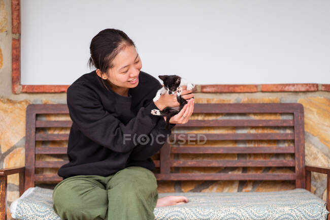 Вміст молодої етнічної жінки, що взаємодіє з чарівною кошеням, сидячи з схрещеними ногами на лавці — стокове фото