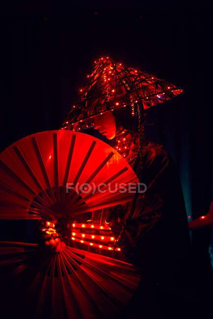 Femme en tenue authentique et chapeau traditionnel avec des lampes lumineuses debout dans un studio sombre avec ventilateur à la main sur fond noir — Photo de stock