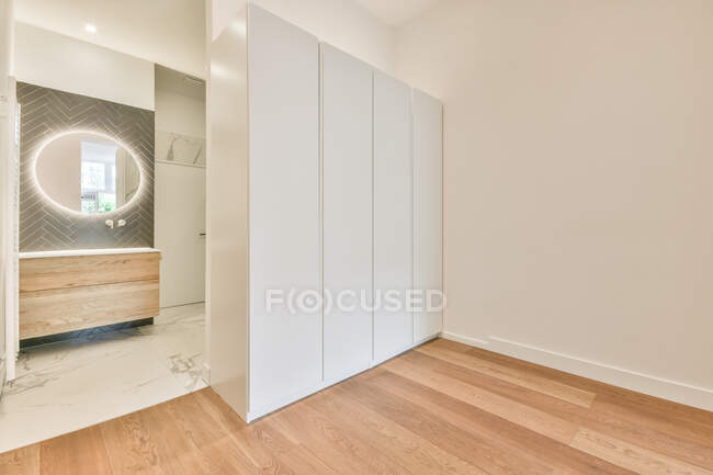 Intérieur de la chambre spacieuse moderne avec garde-robe blanche placée près de la porte de la salle de bain privée avec miroir ovale éclairé et meubles en bois — Photo de stock