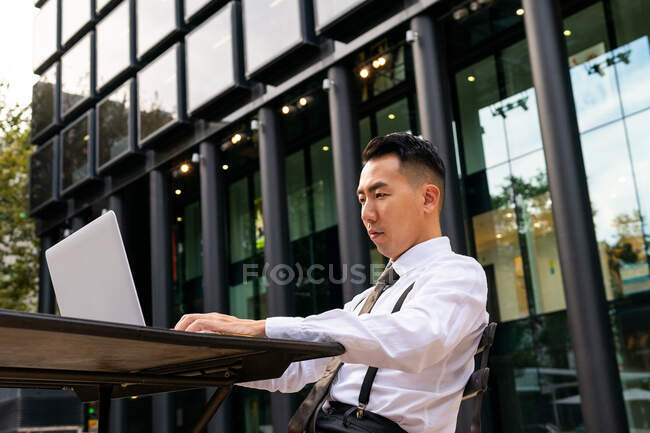 Jeune entrepreneur ethnique surfer sur Internet sur ordinateur portable tout en étant assis à la table de cafétéria urbaine — Photo de stock
