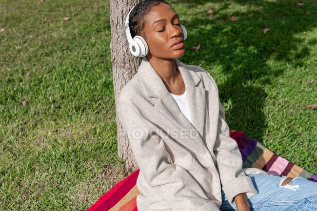 Tranquila mujer afroamericana con los ojos cerrados escuchando música en auriculares inalámbricos mientras está sentada en el césped cerca del tronco del árbol en el soleado parque - foto de stock