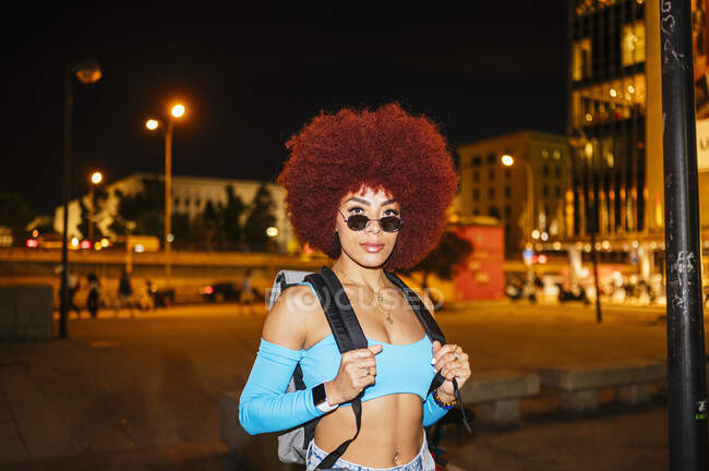 Уверенная женщина с прической афро и модным костюмом с рюкзаком, смотрящая в камеру, стоя на улице со зданиями в вечернее время — стоковое фото