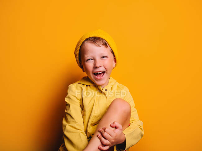 Happy boy in vivid yellow raincoat jacket and beanie hat laughing and looking at camera against yellow background in studio — Stock Photo