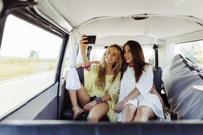 Duas garotas bonitas sentadas dentro de uma van vestida com roupas de verão sorriem enquanto tomam uma selfie — Fotografia de Stock