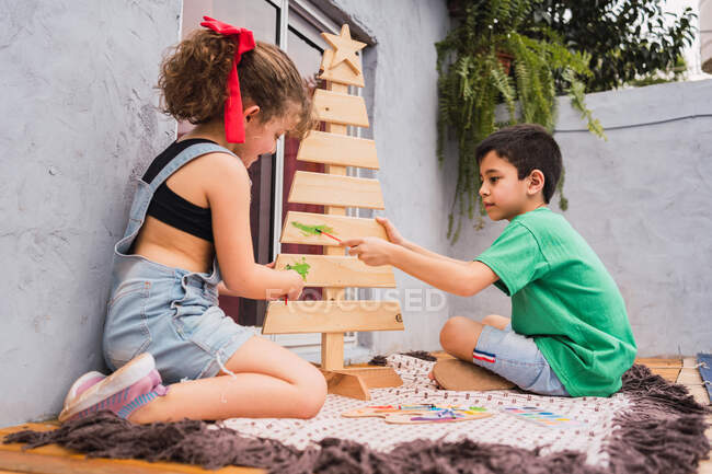 Side view full body of cute kids painting with colorful paints and decorative Christmas tree during holiday preparation in room — Stock Photo