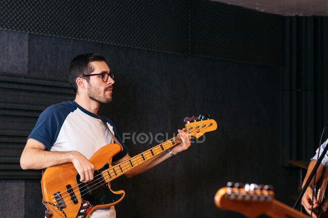 Serious young guy in casual outfit and glasses playing bass guitar in light club — Stock Photo
