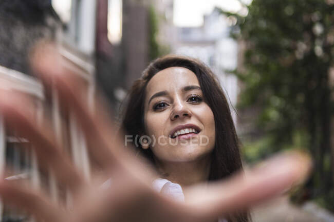 Mujer encantadora tirando de la mano hacia la cámara mientras sonríe y mira a la cámara cerca de los edificios en la calle - foto de stock