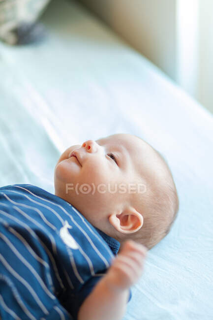 Desde arriba de lindo bebé feliz con el pelo corto en traje de cuerpo rayado sonriendo mientras está acostado en la cama cómoda en la mañana soleada - foto de stock