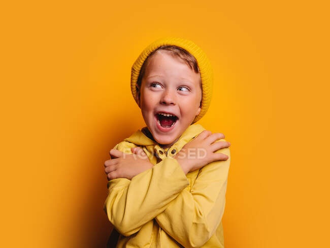 Niño alegre en impermeable de moda y gorro sombrero de pie con los brazos cruzados y mirando hacia otro lado contra el fondo amarillo en el estudio - foto de stock