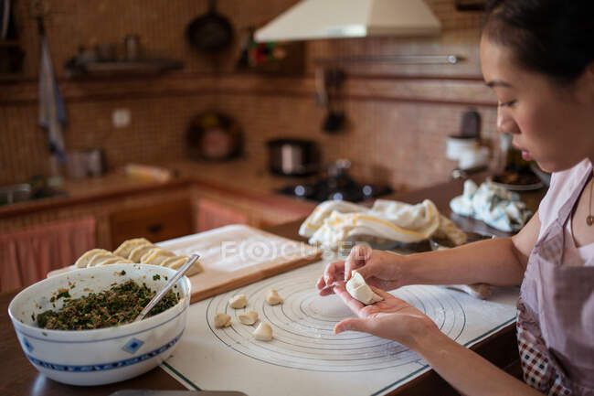 Vista lateral hembra cocinar masa de relleno con carne mientras se preparan albóndigas chinas tradicionales en la cocina - foto de stock