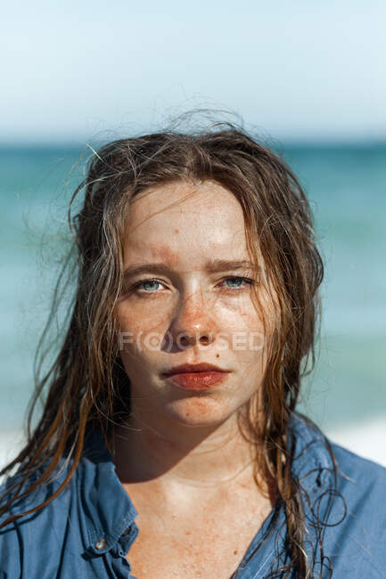 Женщина в мокрой рубашке и с мокрыми волосами, стоящая, глядя на камеру на пляже возле моря, наслаждаясь летним днем — стоковое фото