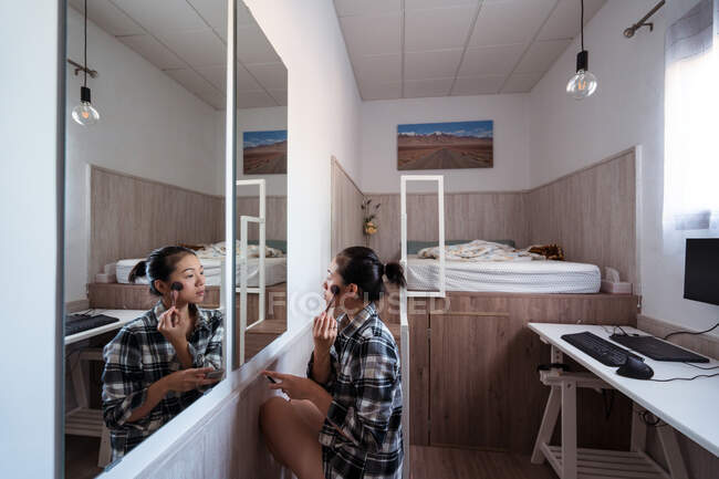 Vue latérale d'une jeune femme ethnique assise près d'un miroir tout en appliquant de la poudre dans une chambre lumineuse avec lit et bureau avec ordinateur — Photo de stock