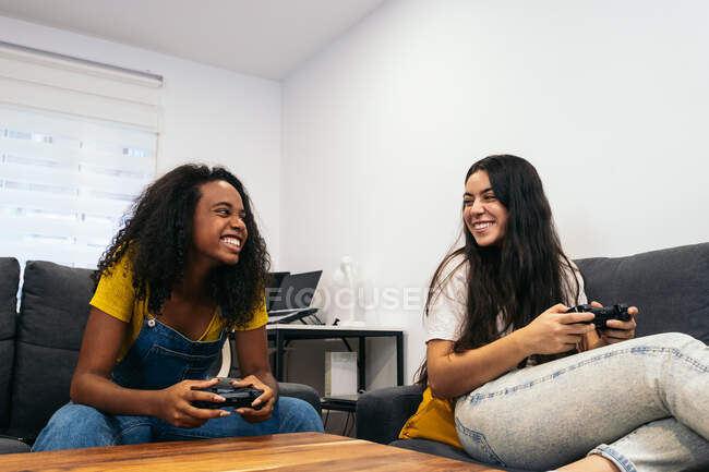 Amis féminines multiraciales positives assis sur le canapé et jouant au jeu vidéo tout en passant du temps ensemble à la maison — Photo de stock