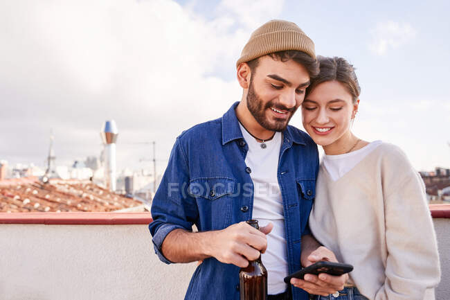 Homem barbudo sorridente com garrafa de cerveja abraçando namorada positiva rolando telefone celular na varanda em dia ensolarado — Fotografia de Stock