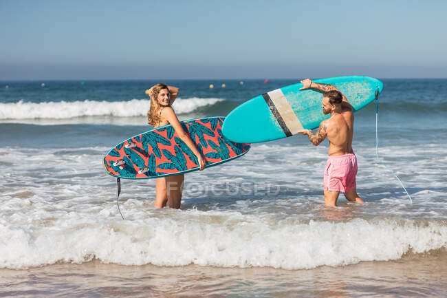 Vista laterale del corpo pieno di coppia sportiva in costume da bagno con tavole da surf che si preparano a navigare in mare ondulato durante l'allenamento oceano ionico — Foto stock