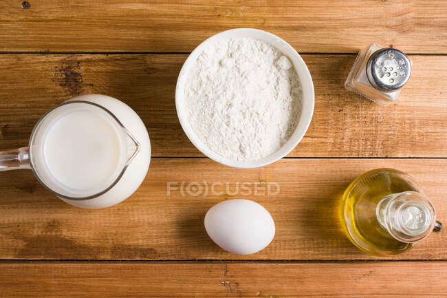 Верхняя чаша с мукой и кувшин с молоком помещены на деревянный стол с яйцом и маслом в светлой кухне — стоковое фото