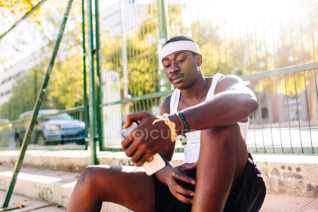 Jugador de baloncesto masculino afroamericano sentado en un campo de deportes y navegando por un teléfono inteligente en un día soleado en verano - foto de stock