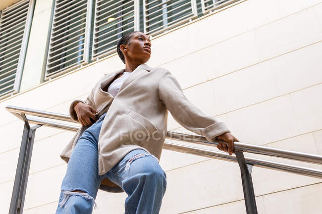 Знизу упевнена афро - американська жінка в модному одязі стоїть на вулиці біля будинку з металевими поручнями в місті. — стокове фото