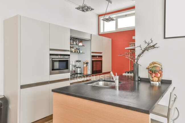Inseltheke mit Unterputzspüle in stilvoller Küche mit verchromten Einbaugeräten in weißem Schrank in heller Wohnung — Stockfoto