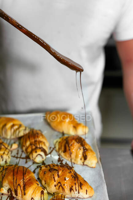 Crop man baker in casual clothes standing and pouring sweet syrup on fresh baked croissants in kitchen — Stock Photo