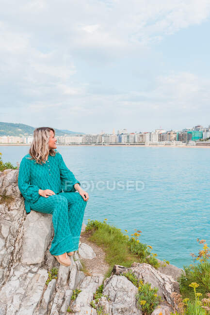 Full body of positive female in stylish outfit sitting on rocks with green plants in San Sebastian in Spain against cloudy blue sky in daytime close to the sea — Stock Photo