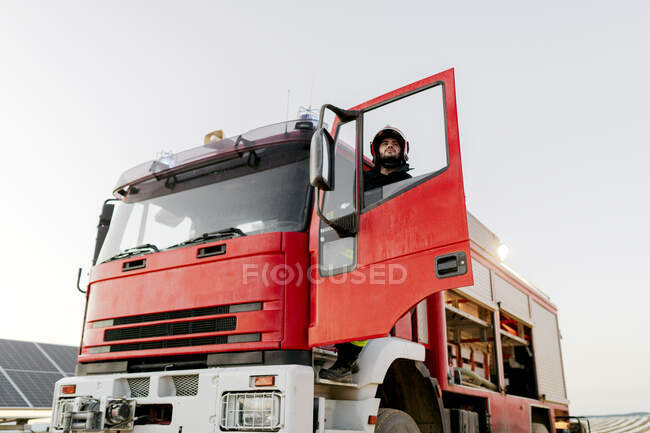 Motorista de bombeiro vestindo uniforme de proteção subindo caminhão de bombeiros enquanto olha para longe — Fotografia de Stock