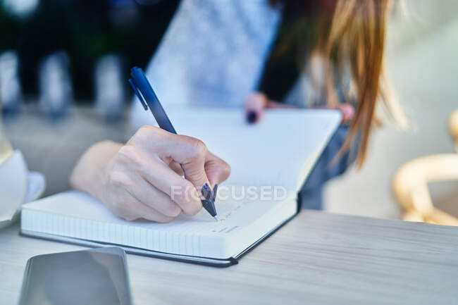 Through glass anonymous female entrepreneur sitting at table with smartphone and writing work related notes in notepad — Stock Photo