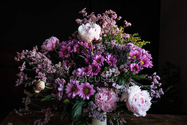 Ramo de peonías frescas y coloridas y crisantemos en jarrón blanco colocado sobre mesa de madera en habitación oscura - foto de stock