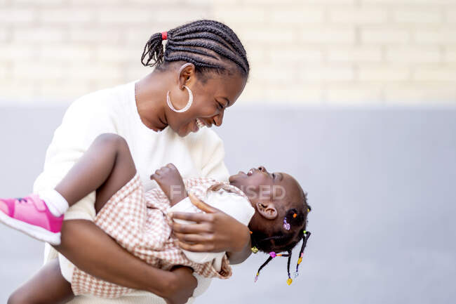 Fröhliche Afroamerikanerin mit Frisur steht bei Tageslicht mit positiver kleiner Tochter im Arm vor Backsteinmauer auf der Straße — Stockfoto
