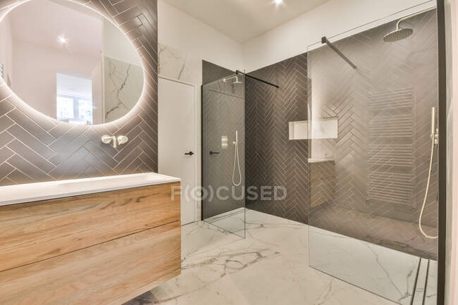 Простора скляна душова кабіна та освітлене овальне дзеркало, що звисає на стіні над раковиною у просторій сучасній ванній кімнаті з мармуровою плиткою — стокове фото