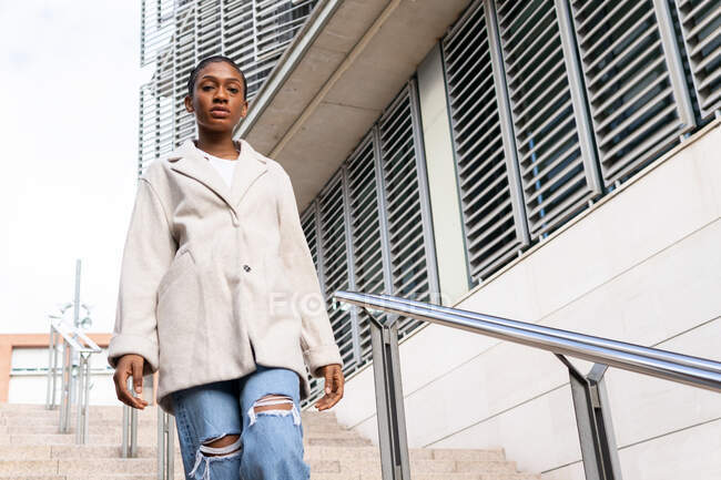 From below of confident African American female in trendy outfit standing on street near building with metal railing in city while walking down the stairs — Stock Photo
