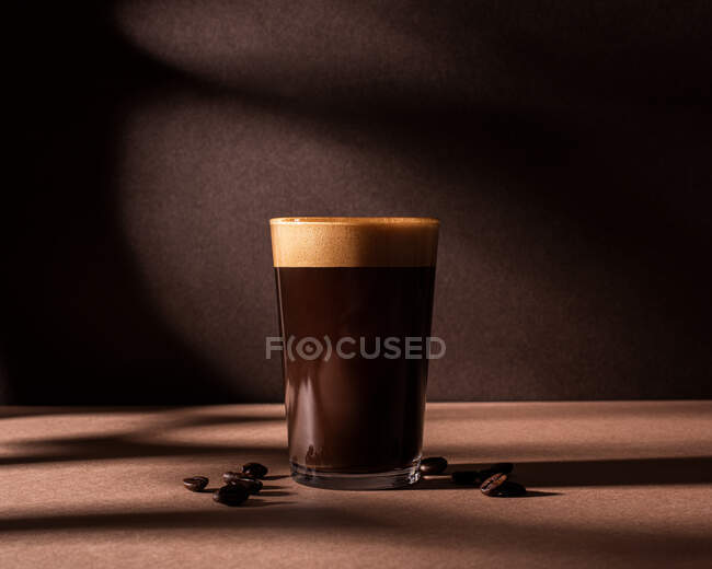 Vorderseite einer schwarzen Kaffeetasse mit einer Schicht Schaum, die neben ein paar Kaffeebohnen auf einem braunen Tisch und einer Wand mit dunklen Schatten steht — Stockfoto