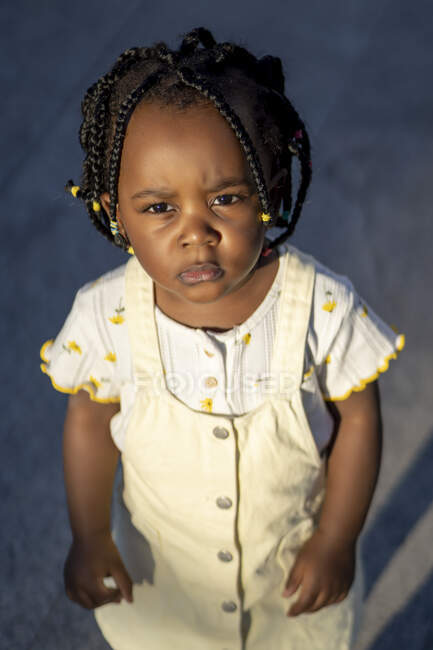 Афроамериканская маленькая девочка с косичками в стильной одежде, стоящая на улице напротив здания в солнечный день — стоковое фото