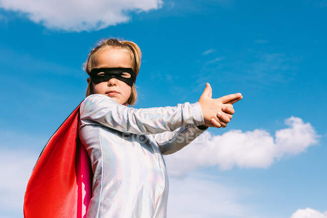 Vista lateral de ángulo bajo de chica confiada en traje de héroe que muestra gesto de pistola para proteger el mundo mientras está de pie contra el cielo - foto de stock