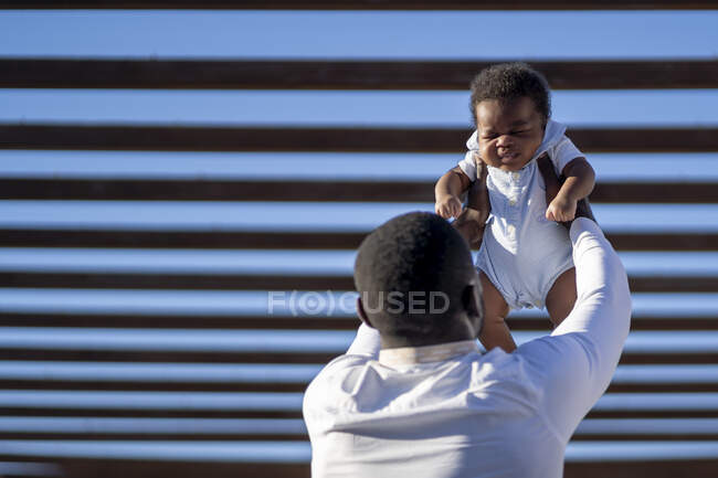 Обратный вид счастливого афроамериканского отца, поднимающего маленького ребенка на руки к голубой стене на улице в солнечный день — стоковое фото
