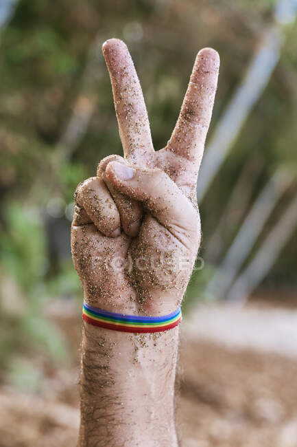 Crop unrecognizable male with rainbow bracelet demonstrating peace gesture against green plants on So Tom and Prncipe island in daylight — Stock Photo