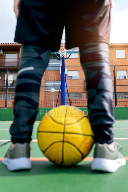 Crop anônimo desportista em activewear de pé em campo de esportes públicos com bola amarela e basquete aro durante o jogo na rua — Fotografia de Stock