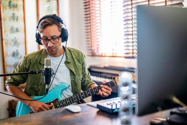 Guitariste masculin avec lunettes et écouteurs tenant l'instrument tout en utilisant l'ordinateur dans le studio moderne — Photo de stock