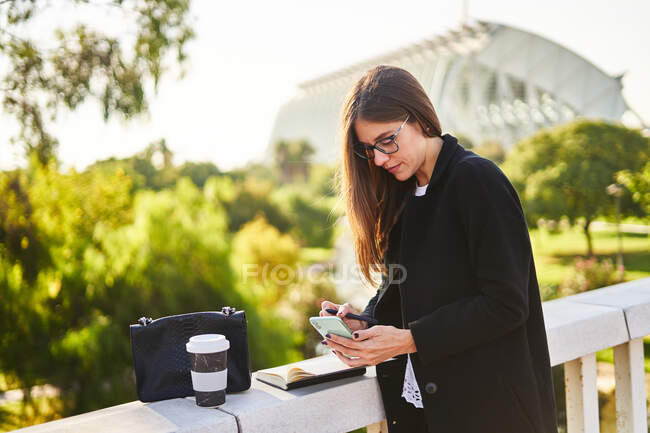 Concentrated businesswoman with brown hair in stylish clothes standing near concrete fence with cup of takeaway coffee and bag and whiting notes in notebook on street in sunny day — Stock Photo