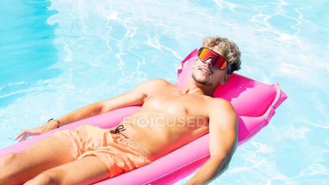 Ruhiger Mann in Badehose und Sonnenbrille sonnt sich an sonnigen Sommertagen auf rosa aufblasbarer Matratze im Schwimmbad mit klarem Wasser — Stockfoto