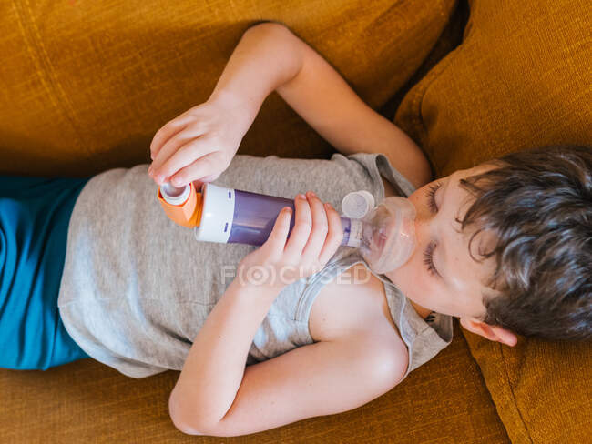 Top view of little boy with asthma using inhaler while lying down on sofa at home — Stock Photo
