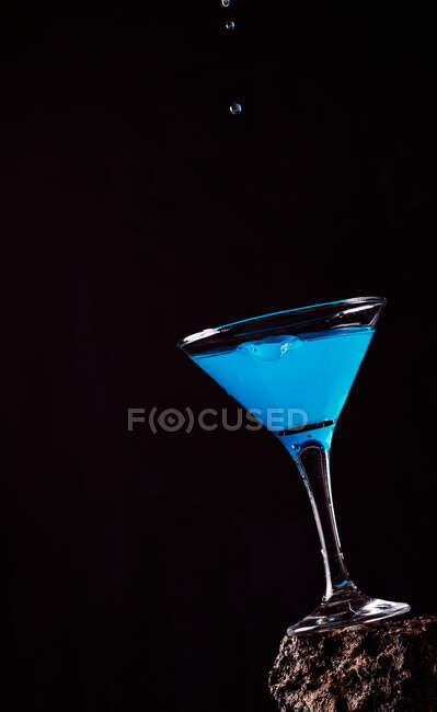 Gocce di cocktail blu Laguna versando in cristallo elegante vetro posto su superficie ruvida su sfondo nero — Foto stock