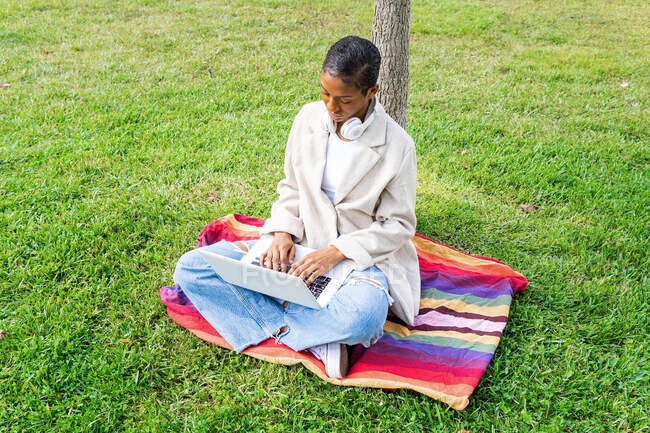 Corps complet de femme afro-américaine concentrée dans les écouteurs tapant sur netbook moderne tout en étant assis sur la pelouse herbeuse dans le parc — Photo de stock