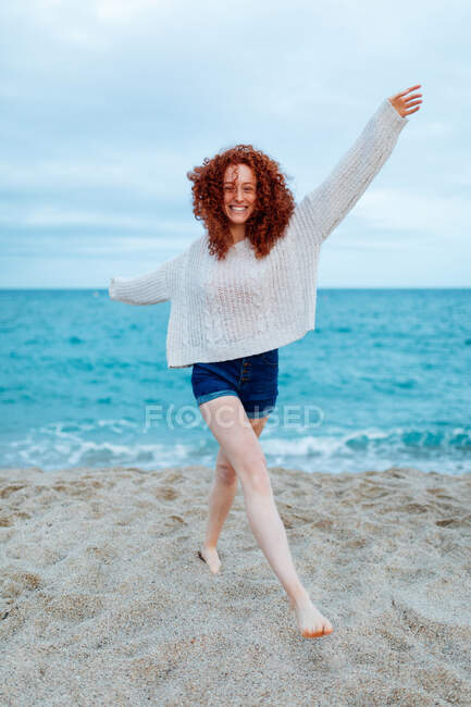 Full length of barefoot smiling female traveler standing looking at camera on sandy coast washed by foamy waves of blue sea — Stock Photo