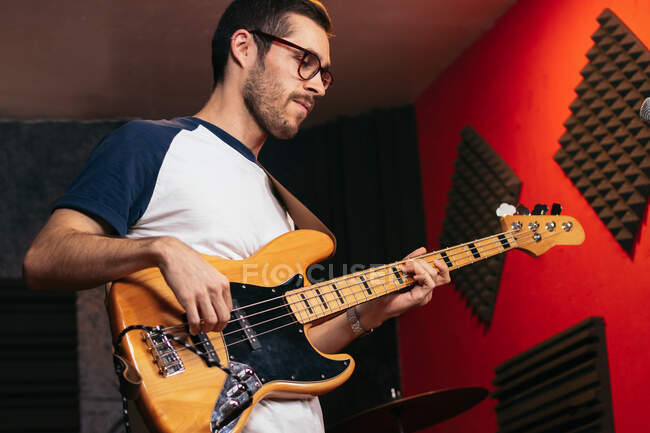 Serious young guy in casual outfit and glasses playing bass guitar in light club — Stock Photo