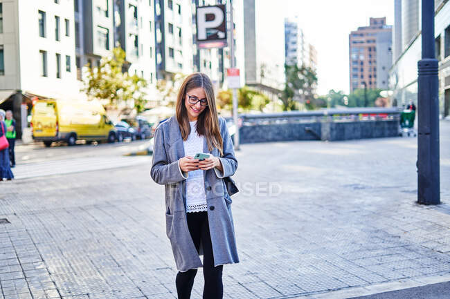 Donna positiva in abbigliamento formale utilizzando il cellulare mentre cammina sulla strada della città al lavoro — Foto stock